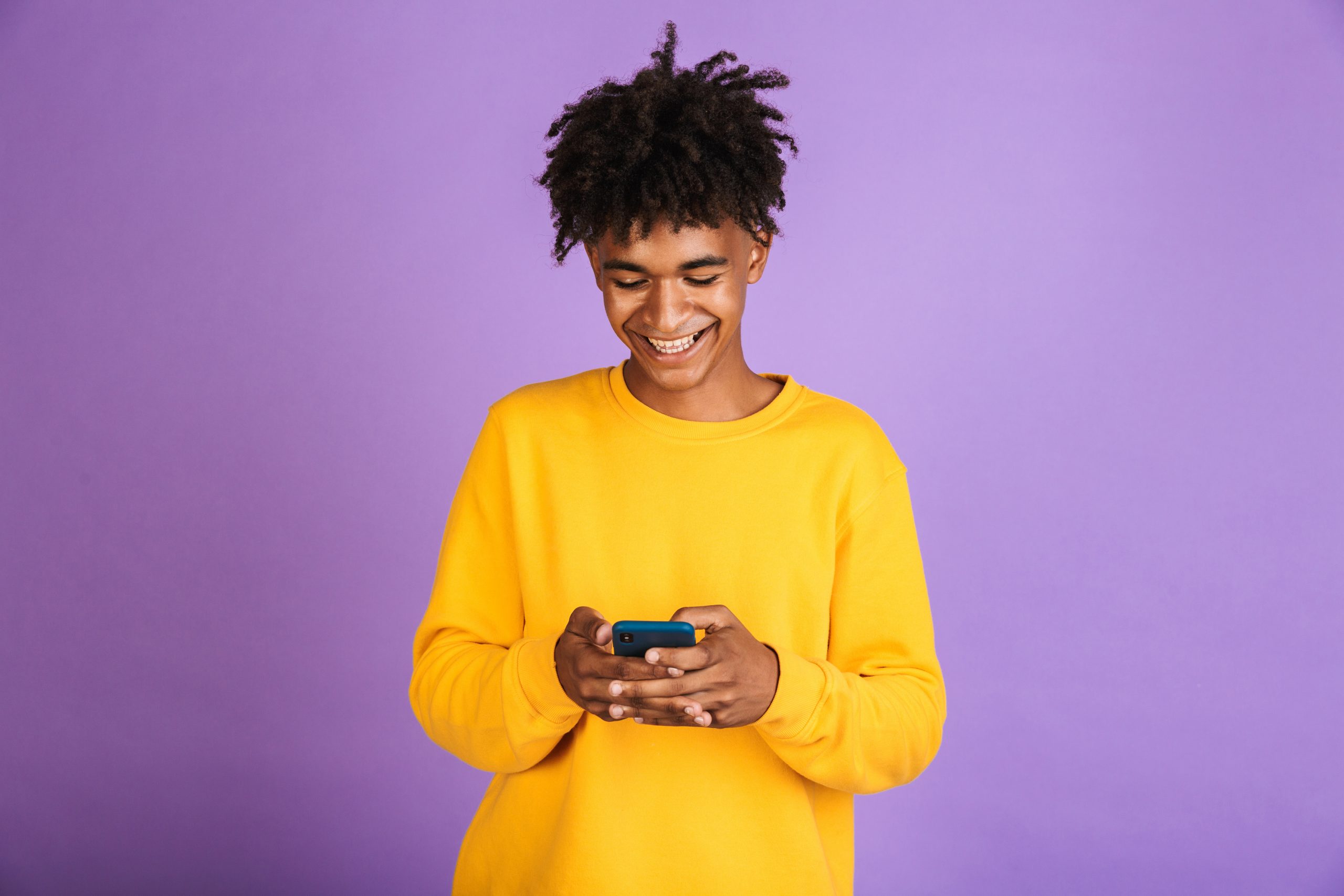 Portrait of stylish american guy smiling and holding smartphone, isolated over violet background